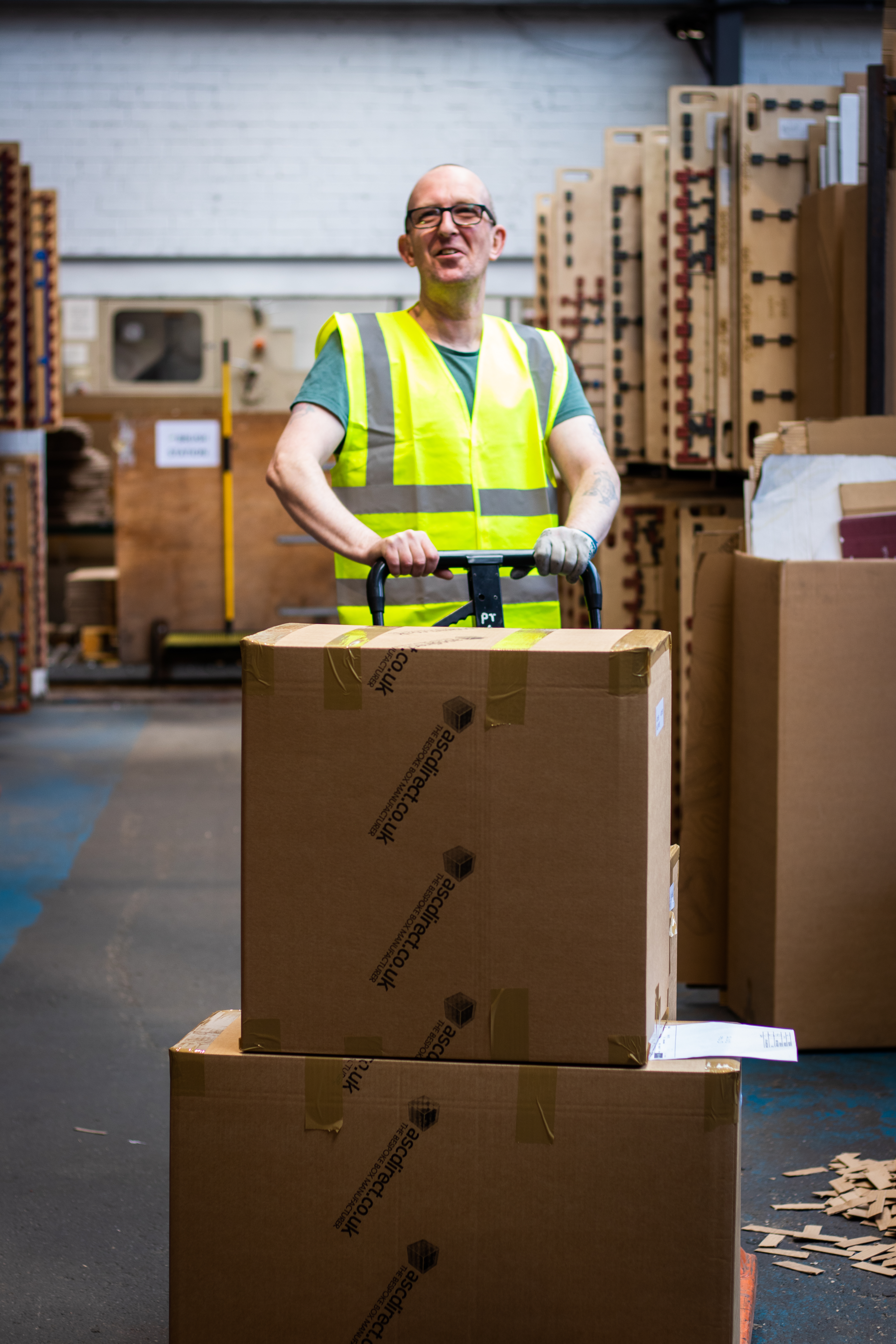 Smiling white male warehouse staff member pushing ascdirect.co.uk labelled boxes on a trolley